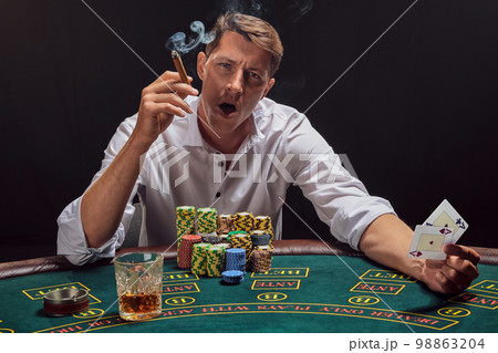 Handsome emotional man is playing poker sitting at the table in casino against a white spotlight. 98863204