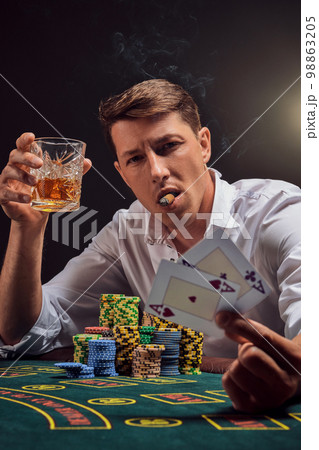 Handsome emotional man is playing poker sitting at the table in casino against a white spotlight. Handsome emotional man is playing poker sitting at the table in casino against a white spotlight. 98863205