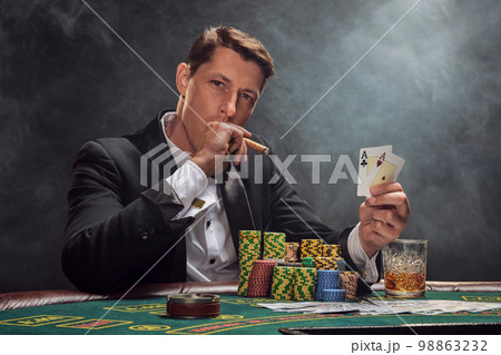 Handsome emotional man is playing poker sitting at the table in casino against a white spotlight. Handsome emotional man is playing poker sitting at the table in casino against a white spotlight. 98863232