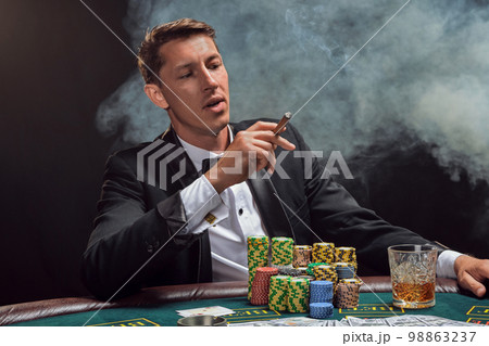 Handsome emotional man is playing poker sitting at the table in casino against a white spotlight. 98863237
