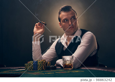 Wealthy man is smoking a cigar and playing poker with an excitement at a casino on black background. Wealthy man is smoking a cigar and playing poker with an excitement at a casino on black background. 98863242