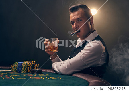 Wealthy man is smoking a cigar and playing poker with an excitement at a casino on black background. Wealthy man is smoking a cigar and playing poker with an excitement at a casino on black background. 98863243