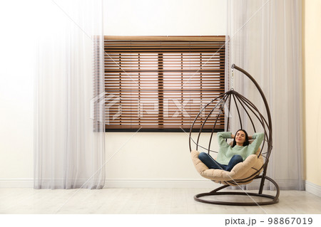 Young woman relaxing in hanging chair near window at home. Space for text Young woman relaxing in hanging chair near window at home. Space for text 98867019