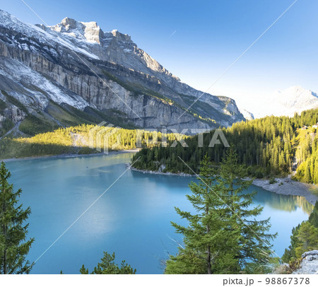 Idyllic morning view of the lake Oeschinensee. Location Swiss alps, Switzerland, Kandersteg district. Blue mountain lake with pine trees and mountains in background. Idyllic morning view of the lake Oeschinensee. Location Swiss alps, Switzerland, Kandersteg district. Blue mountain lake with pine trees and mountains in background. 98867378