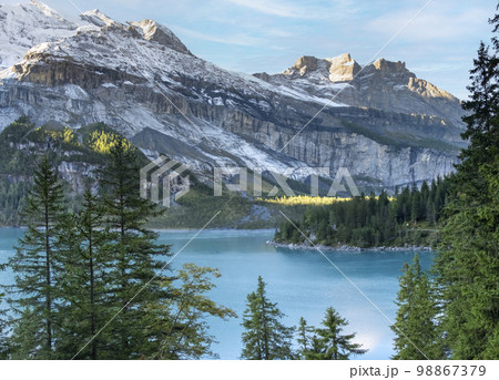 Idyllic morning view of the lake Oeschinensee. Location Swiss alps, Switzerland, Kandersteg district. Blue mountain lake with pine trees and mountains in background. Idyllic morning view of the lake Oeschinensee. Location Swiss alps, Switzerland, Kandersteg district. Blue mountain lake with pine trees and mountains in background. 98867379
