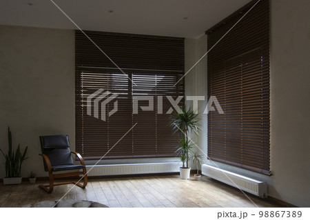 Wooden blinds on large windows in the interior. Living room with armchair and houseplants near windows with wood blinds. Motorized jalousie in the smart house. Wooden blinds on large windows in the interior. Living room with armchair and houseplants near windows with wood blinds. Motorized jalousie in the smart house. 98867389