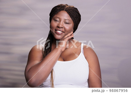 Curvy woman touching smooth hydrated skin after skincare procedures and looking at camera. Plus size african american model holding palm on face after cosmetology treatment 98867956