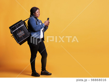 Full body of pizzeria courier carrying takeaway thermal backpack while checking client adreess on smartphone before start deliver food order during lunch time. Takeout service concept Full body of pizzeria courier carrying takeaway thermal backpack while checking client adreess on smartphone before start deliver food order during lunch time. Takeout service concept 98868016