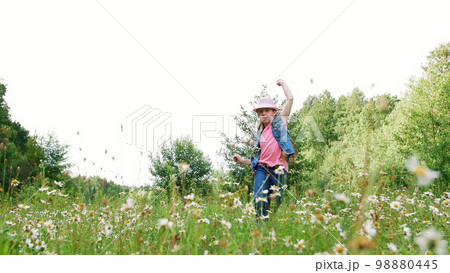 In the grass, among the daisies, in the meadow, dancing, jumping, having fun, a pretty girl, about seven years old. She has long blond hair, is dressed in a denim vest and a pink hat In the grass, among the daisies, in the meadow, dancing, jumping, having fun, a pretty girl, about seven years old. She has long blond hair, is dressed in a denim vest and a pink hat 98880445