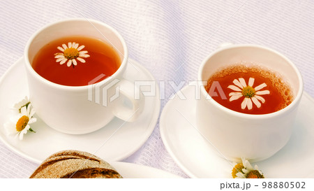 A close-up, a white plate with pastries, biscuits and cookies, decorated with white daisies. On white saucers there are two white cups with chamomile tea A close-up, a white plate with pastries, biscuits and cookies, decorated with white daisies. On white saucers there are two white cups with chamomile tea 98880502
