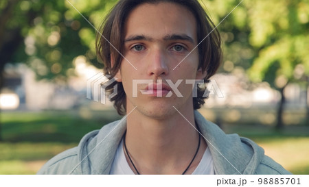 Close up face of an young man is looking to the camera,  in the street. Man with calm emotions on his face looks into the camera - outdoors. Handsome guy posing outdoors on green trees background. 98885701