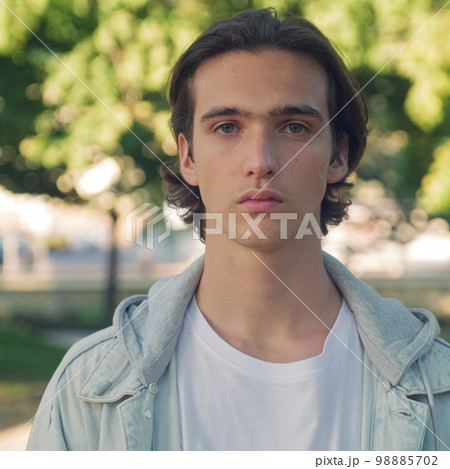 Close up face of an young man is looking to the camera,  in the street. Man with calm emotions on his face looks into the camera - outdoors. Handsome guy posing outdoors on green trees background. 98885702