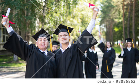 A group of graduates in robes outdoors. An elderly man and a young guy congratulate each other on receiving a diploma. A group of graduates in robes outdoors. An elderly man and a young guy congratulate each other on receiving a diploma. 98886862