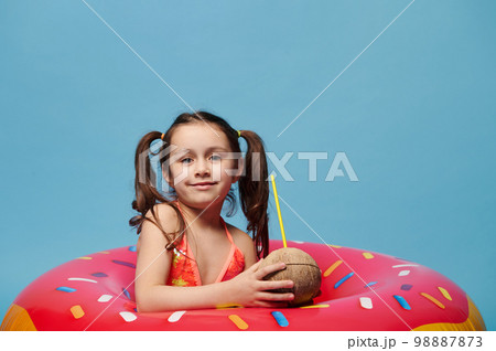Cute child girl with coconut cocktail smiles looking at camera, posing inside a donut swim ring on blue color background Cute child girl with coconut cocktail smiles looking at camera, posing inside a donut swim ring on blue color background 98887873