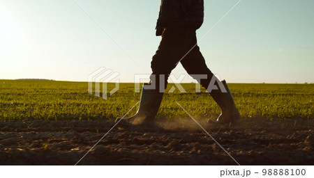 Farmer walks in rubber boots  down a farmer field  dust rising from shoes. Low angle. One part is sown, the second part is not sown. 98888100