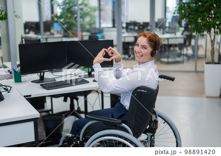 Caucasian woman in wheelchair showing heart sign while sitting in office. Female call center worker. Caucasian woman in wheelchair showing heart sign while sitting in office. Female call center worker. 98891420
