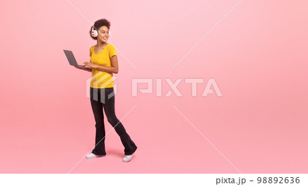 Cheerful young african american curly woman in casual and wireless headphones goes with laptop Cheerful young african american curly woman in casual and wireless headphones goes with laptop 98892636
