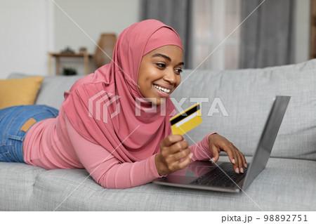Online shopping. Positive black lady in hijab with laptop and credit card lying on sofa, buying goods in web store 98892751