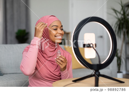 Happy black muslim female beauty blogger making video on smartphone set on ring lamp, showing new facial serum 98892786