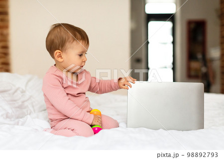 Cute little baby girl sitting on bed and touching laptop computer, bedroom interior, free copy space 98892793