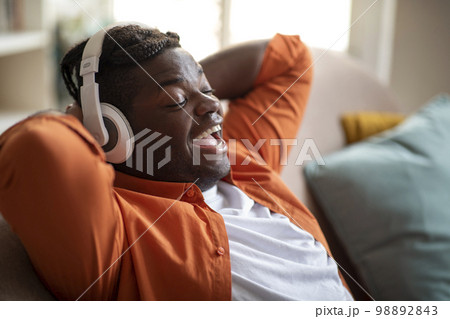 Joyful black guy reclining on sofa, singing songs, using headphones 98892843