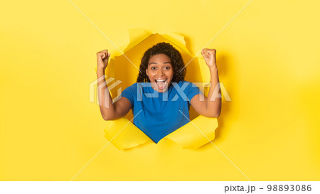 Overjoyed black woman cheering with clenched fists, celebrating victory, posing in torn hole of yellow background Overjoyed black woman cheering with clenched fists, celebrating victory, posing in torn hole of yellow background 98893086