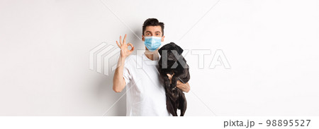 Covid-19, animals and quarantine concept. Young man in medical mask holding cute black pug dog, showing okay sign, like and approve, standing over white background 98895527