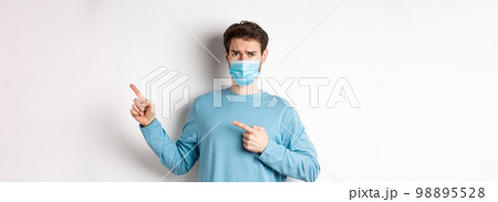 Coronavirus, health and quarantine concept. Sad man in medical mask frowning, pointing fingers right with doubtful face, standing over white background 98895528