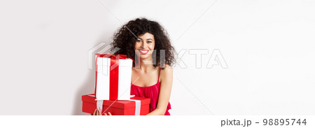 Beautiful birthday girl with curly hair, holding bday gifts and smiling happy, celebrating, standing against white background 98895744