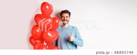 Smiling caucasian man standing with heart balloon, prepare surprise for lover on Valentines day, showing thumbs up and looking at camera, white background 98895746