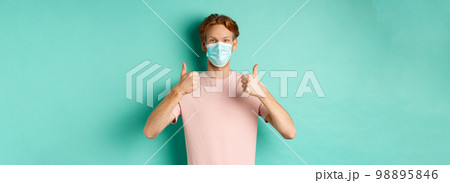 Covid-19, pandemic and lifestyle concept. Cheerful redhead guy in medical mask showing thumbs up in approval, like and praise product, standing over turquoise background Covid-19, pandemic and lifestyle concept. Cheerful redhead guy in medical mask showing thumbs up in approval, like and praise product, standing over turquoise background 98895846
