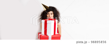 Cute birthday girl with curly hair and party hat, holding gifts and looking happy at camera, standing against white background Cute birthday girl with curly hair and party hat, holding gifts and looking happy at camera, standing against white background 98895856