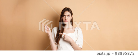 Hesitant young woman look unsure, pointing fingers right while making decision, frowning with puzzled face, standing on beige background 98895875