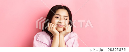 Close-up portrait of dreamy and romantic asian girl, lean face on hands and smiling, looking with admiration and happiness, standing against pink background 98895876
