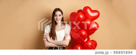 Cheerful young woman looking happy on Valentines day, standing near hearts balloons with arms crossed, smiling at camera, standing on beige background 98895894