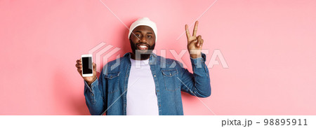 Online shopping and technology concept. Happy Black guy in beanie and denim shirt showing mobile screen and peace sign, standing over pink background Online shopping and technology concept. Happy Black guy in beanie and denim shirt showing mobile screen and peace sign, standing over pink background 98895911