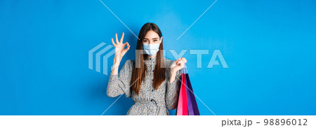 Covid-19, pandemic and lifestyle concept. Attractive woman wear medical mask on shopping, show okay and hold bags with purchases, blue background 98896012