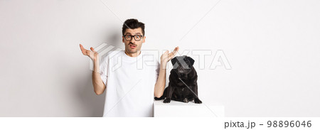 Image of confused man in glasses raising hands and shrugging complicated, standing near black pug dog over white background Image of confused man in glasses raising hands and shrugging complicated, standing near black pug dog over white background 98896046