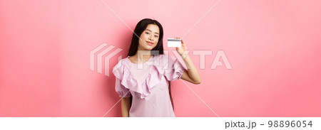 Shopping. Young korean girl in dress showing credit card, smiling satisfied at camera, standing on pink background Shopping. Young korean girl in dress showing credit card, smiling satisfied at camera, standing on pink background 98896054
