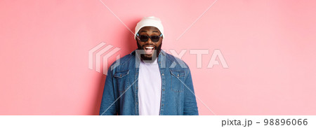 Surprised and happy african-american hipster man in beanie and sunglasses, smiling and looking at camera, standing confident over pink background 98896066