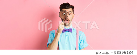 Funny man in bow-tie look through magnifying glass, squinting and making silly faces, standing on pink background 98896134