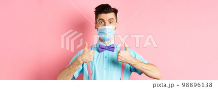 Coronavirus, healthcare and quarantine concept. Satisfied guy in medical mask showing thumbs up in approval, praising excellent choice, pink background 98896138