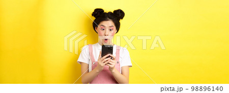 Asian teen girl look excited at smartphone screen, reading news on phone, standing on yellow background Asian teen girl look excited at smartphone screen, reading news on phone, standing on yellow background 98896140