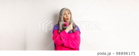 Curious asian senior female checking out promo, looking and pointing finger left, standing over white background 98896175