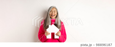 Real estate. Happy asian grandmother laughing with eyes closed, holding paper house model, dreaming about paper house model, standing over white background Real estate. Happy asian grandmother laughing with eyes closed, holding paper house model, dreaming about paper house model, standing over white background 98896187