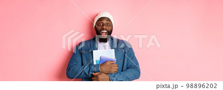 Image of adult african-american man holding notebooks and smiling, studying at courses, standing over pink background Image of adult african-american man holding notebooks and smiling, studying at courses, standing over pink background 98896202
