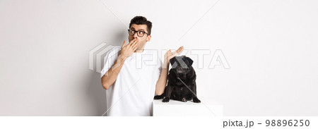 Surprised and shocked man in glasses, standing near cute black dog and staring right at logo, posing over white background 98896250