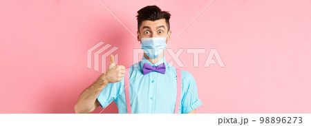 Coronavirus, healthcare and quarantine concept. Impressed young man in medical mask showing thumb up, looking satisfied, recommending good product, pink background 98896273