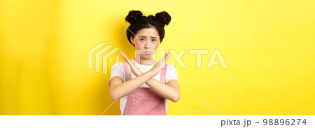 Sad girl begging to stop, frowning uspet and showing cross sign, say no, standing gloomy on yellow background Sad girl begging to stop, frowning uspet and showing cross sign, say no, standing gloomy on yellow background 98896274