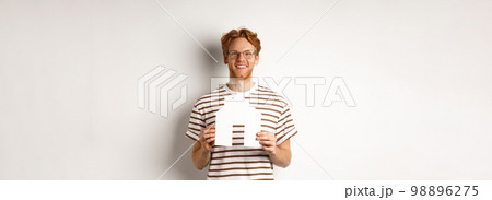 Handsome young man with beard and red hair, wearing glasses and striped t-shirt, showing paper house cutout and smiling, concept of real estate and buying property 98896275